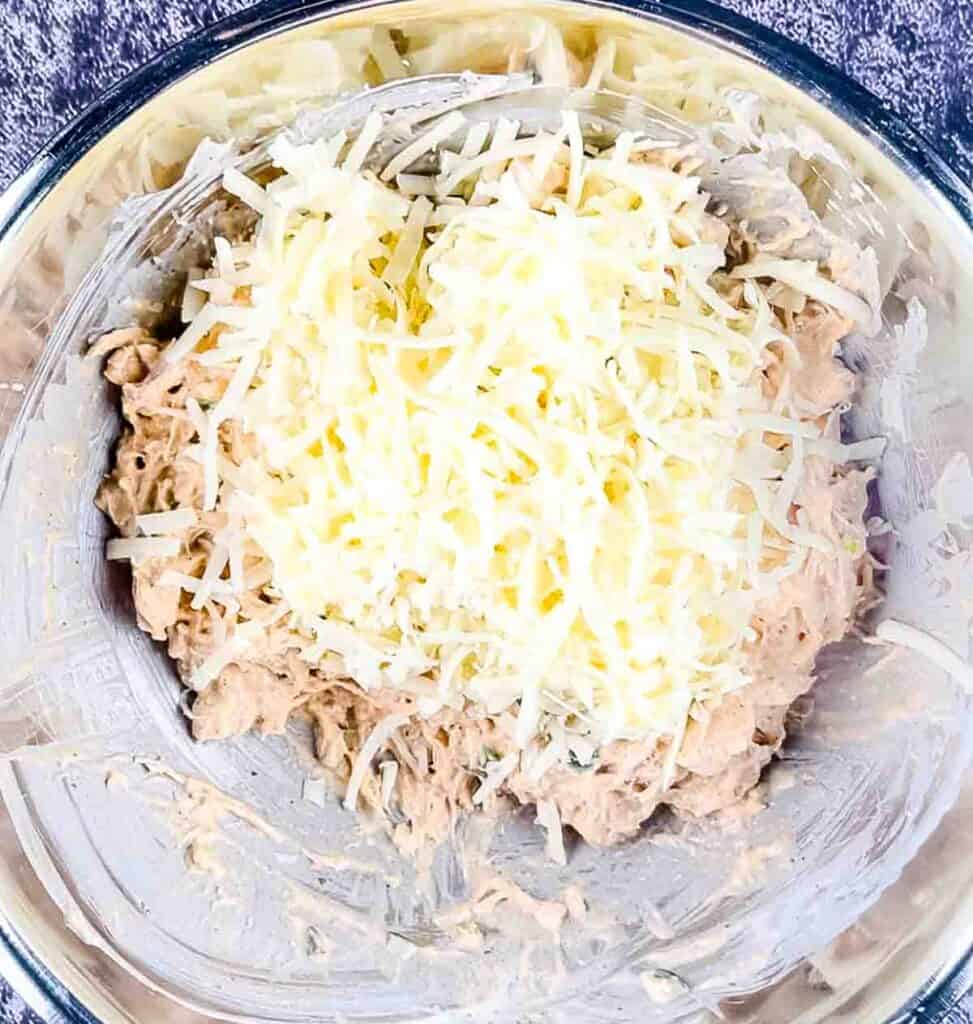 Shredded cheese added to the chicken in a bowl with sour cream, seasoning, hot sauce, and ranch dressing.