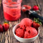 Three scoops of creamy strawberry sorbet in a white bowl with the pint container and an ice cream scoop in the background.