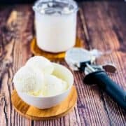 Three scoops of vanilla ice cream in a white bowl with a pint jar and ice cream scoop in the background.
