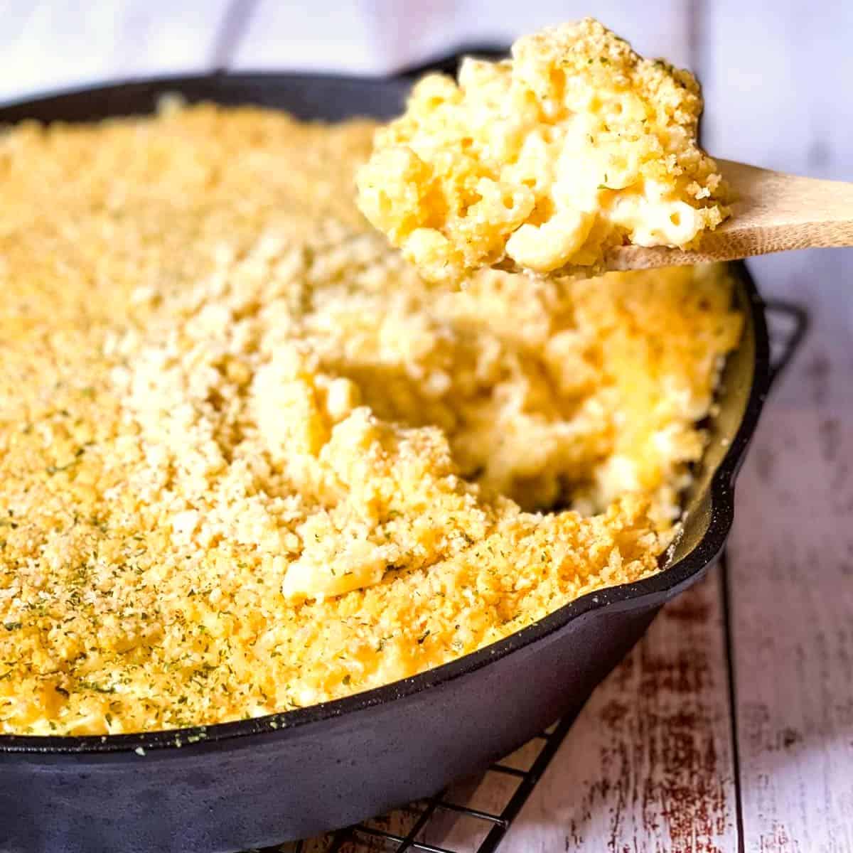 The cast iron pan of mac n cheese with a wooden spoon lifting up a spoonful.