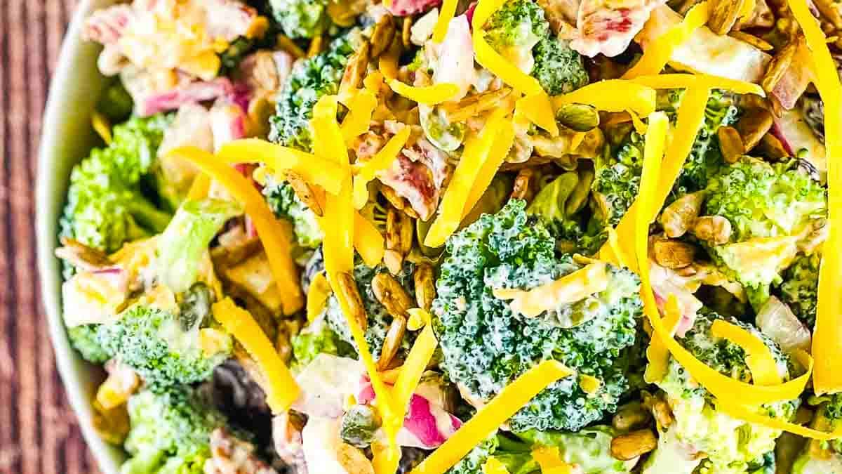 A bowl of Broccoli Crunch Salad with fresh broccoli, crispy bacon, and creamy dressing, garnished with shredded cheese, served in a white dish on a wooden table.