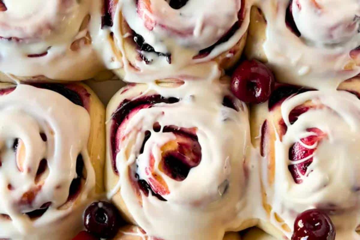 Cherry cinnamon rolls covered in cream cheese frosting, garnished with whole cherries.
