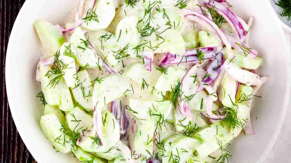 A bowl of creamy cucumber salad with mayo, garnished with fresh herbs, served in a white dish on a wooden table.