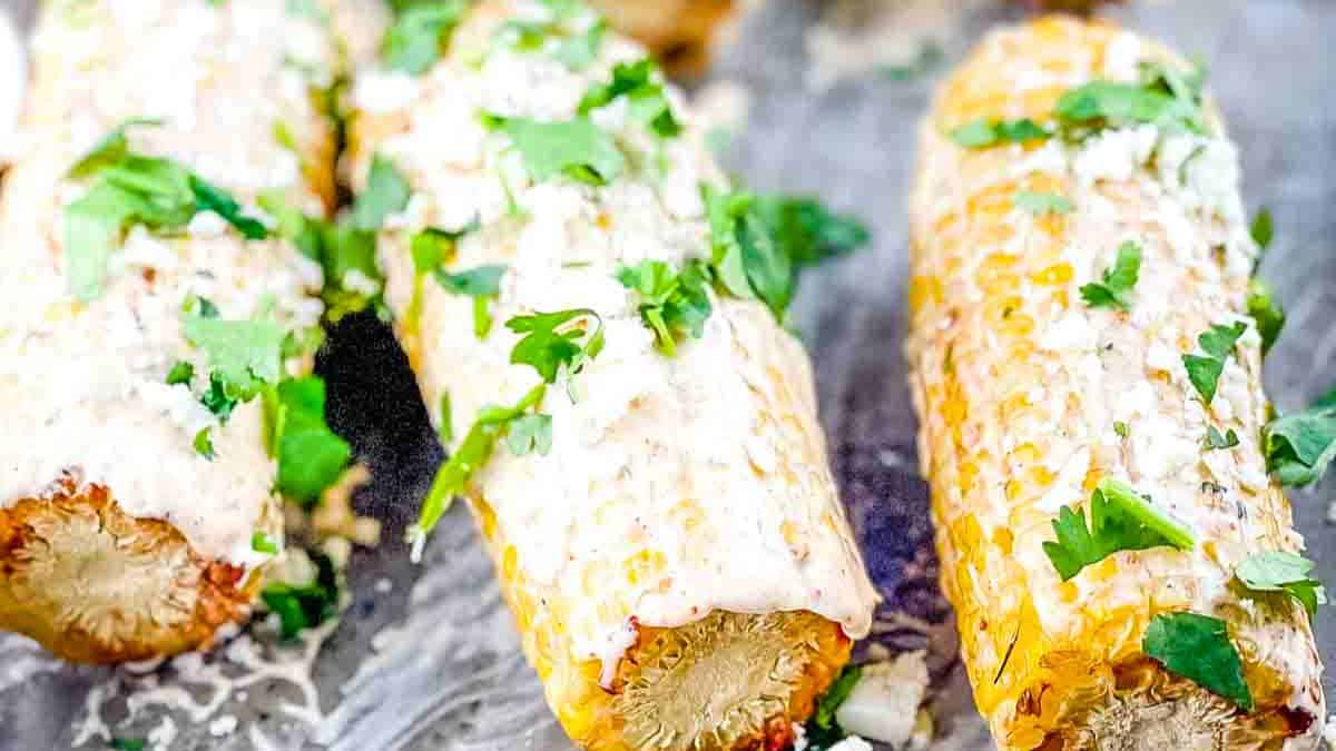 Grilled Mexican Street Corn on the cob, coated with creamy sauce and sprinkled with cheese and spices, served on a rustic cookie sheet.