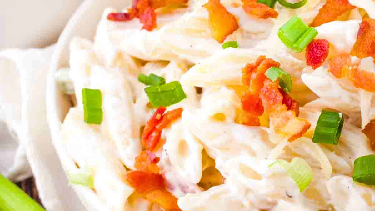 A bowl of Bacon Ranch Pasta Salad with pasta, crispy bacon, and creamy ranch dressing, garnished with fresh herbs, served in a white dish on a rustic table.
