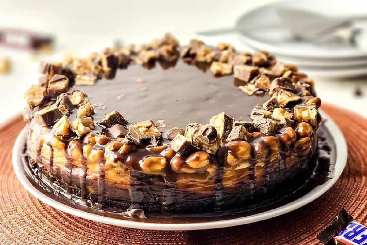 A Snickers Cheesecake on a plate, featuring a creamy filling dotted with chunks of Snickers, drizzled with caramel and chocolate, and sprinkled with peanuts for an extra crunch.