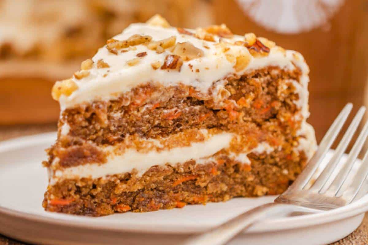 A slice of Carrot Cake inspired by Starbucks, showcasing moist layers of cake with flecks of carrot and nuts, topped with a thick layer of creamy white frosting, served on a simple white plate.