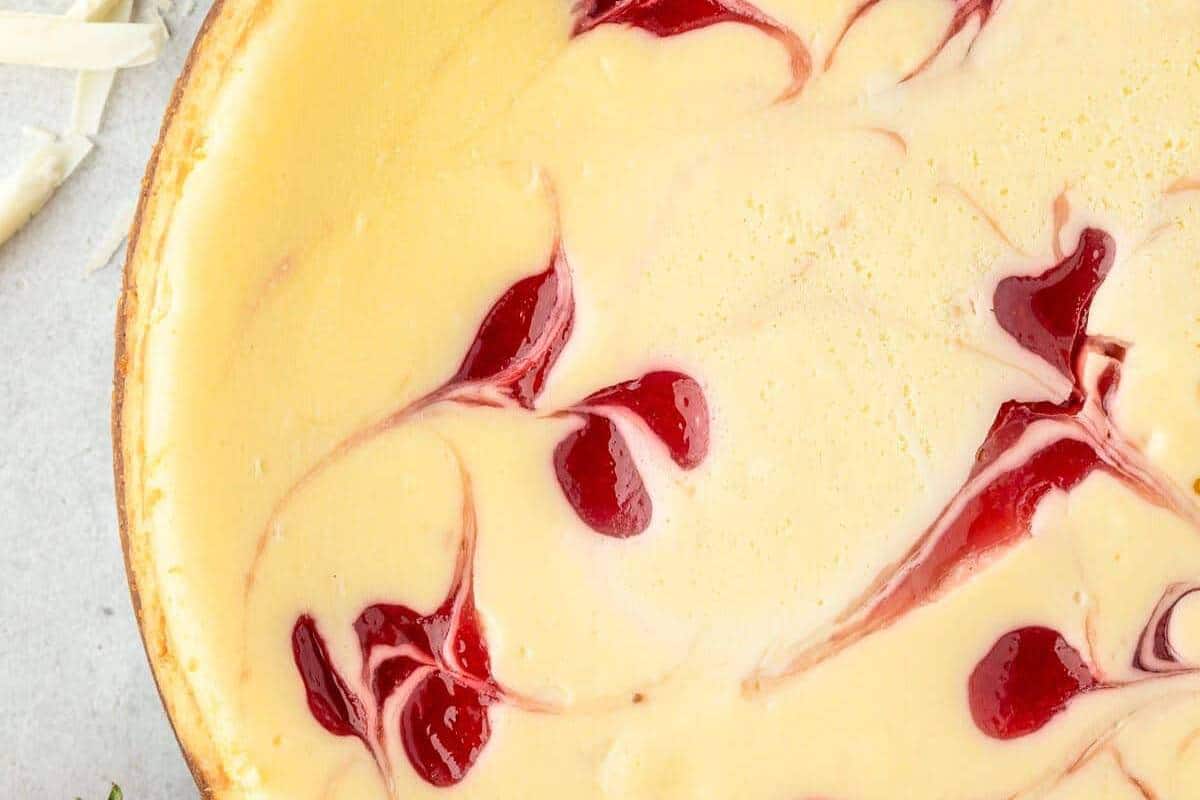 A Strawberry White Chocolate Cheesecake on a plate, showcasing the elegant swirls of bright strawberry sauce mixed into a smooth white chocolate cheesecake base.