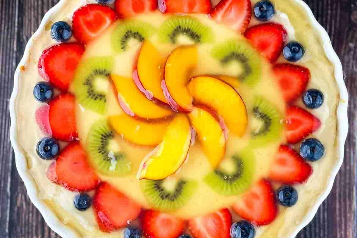 A large, round sugar cookie fruit pizza featuring a smooth layer of cream cheese frosting and topped with an array of fresh fruit slices arranged in a cheerful pattern, perfect for Mother's Day dessert.