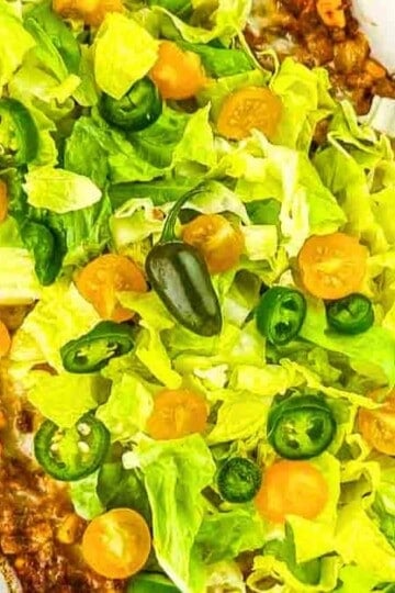 A casserole dish filled with walking taco casserole, topped with chopped lettuce, yellow tomatoes, and sliced jalapeno peppers.