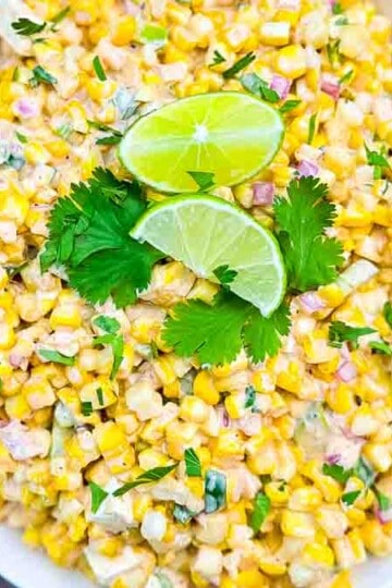 Grilled corn kernels mixed with red onion, jalapeño, cilantro, Cotija cheese, and diced avocado, tossed in a creamy, tangy dressing, served in a white bowl.
