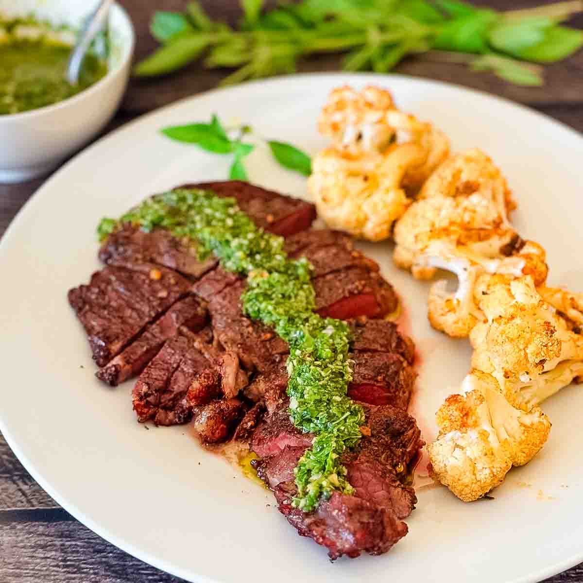 A grilled steak sliced on a dinner plate and topped with green chimichurri sauce.