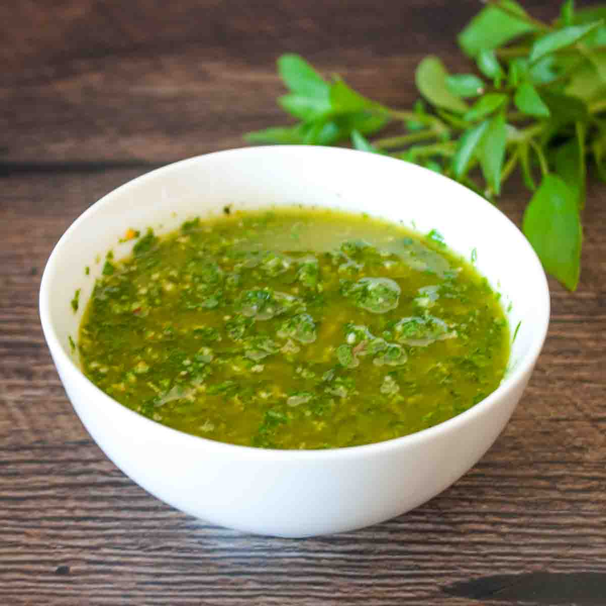 Basil chimichurri in a white serving bowl on a wooden table, with a bunch of fresh Thai basil in the background.