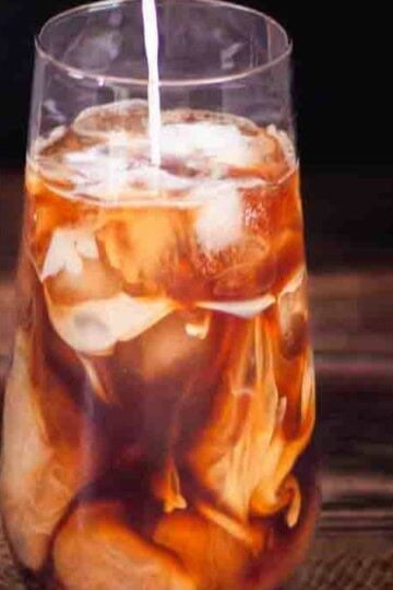 Cream being poured over coffee and ice cubes in a tall glass to make the perfect iced coffee at home.