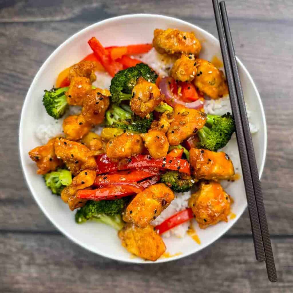 A white bowl filled with rice and honey buffalo chicken with broccoli and peppers, garnished with sesame seeds.