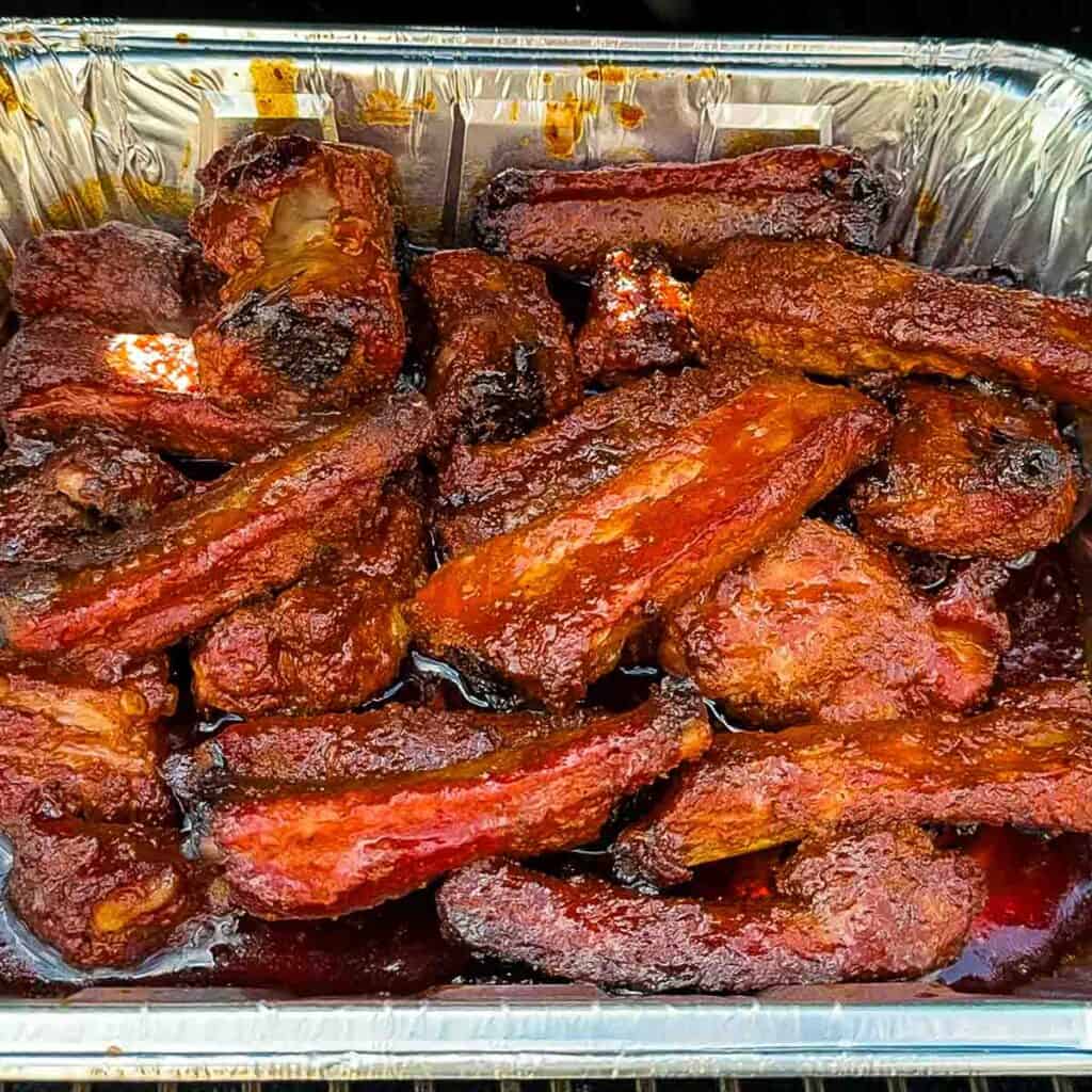 Uncovering ribs in a foil pan on the smoker, showing a caramelized glaze after additional cooking.