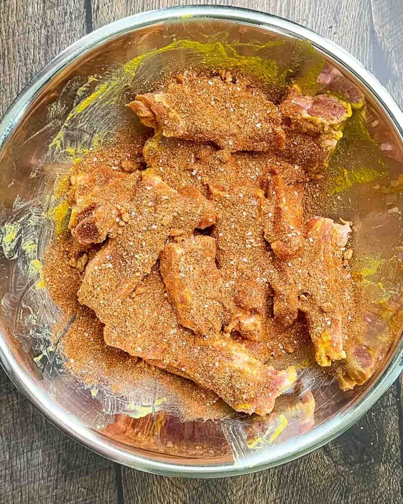 Generously sprinkling pork dry rub over ribs in a bowl, ensuring every inch is covered.