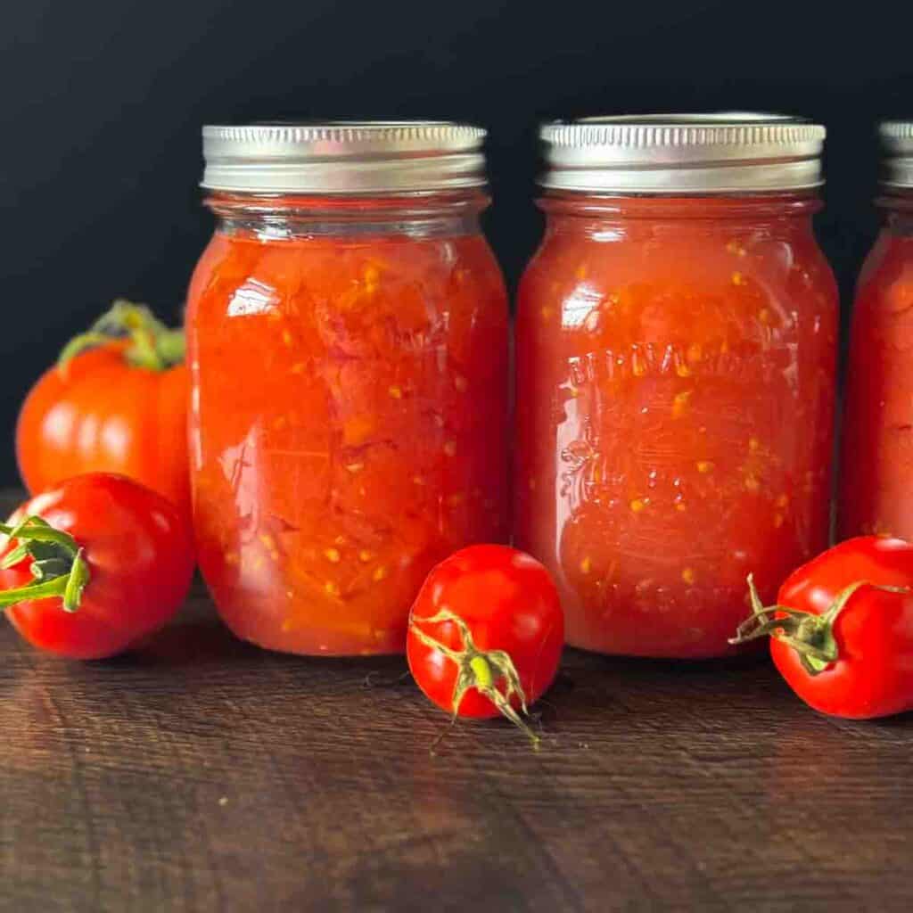 Jars filled with homemade canned tomatoes on a wooden counter with fresh tomatoes beside them.