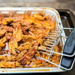 Pulled pork loin in an aluminum pan, pulled apart, with meat hooks sitting on the edge.