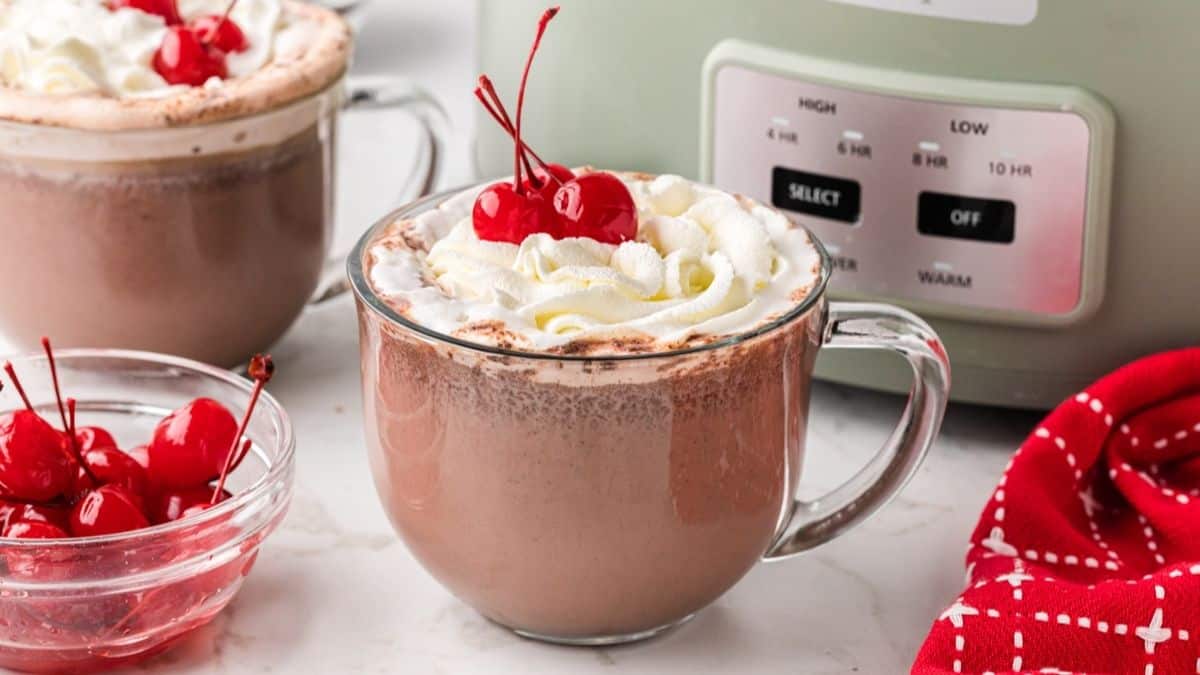 Hot chocolate in a clear mug, topped with whipped cream and cherries.