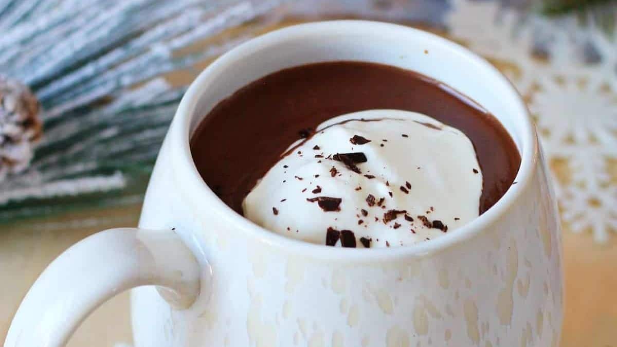 Thick, dark French hot chocolate served in a white mug, topped with a cloud of whipped cream and dusted with cocoa powder, looking rich and decadent.