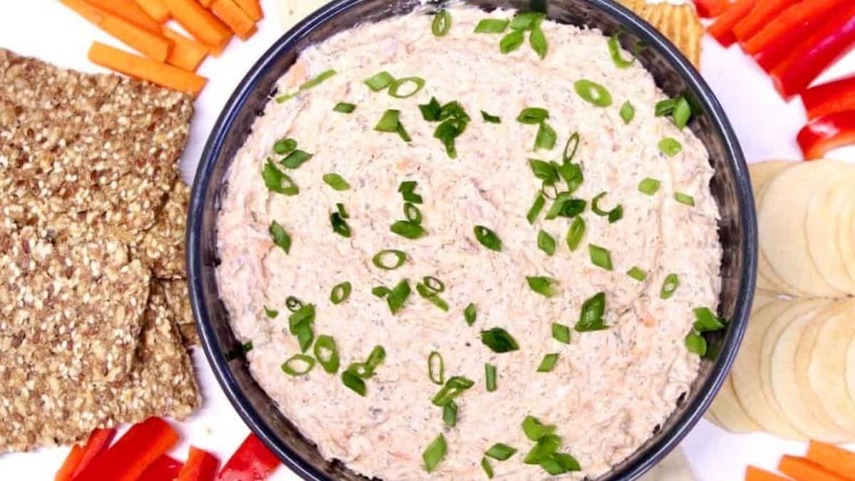 Pink-hued cream cheese dip with chunks of salmon and fresh herbs in a serving bowl surrounded by dippers.