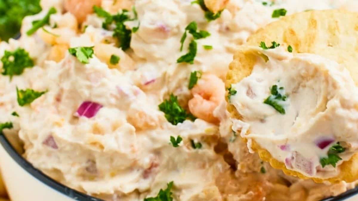 Creamy pink-tinted dip with visible pieces of chopped shrimp and herbs, being scooped up on a cracker.