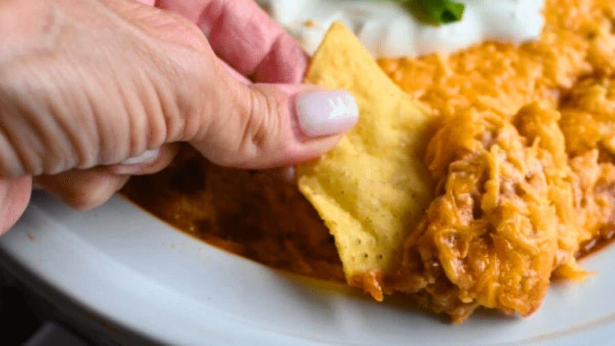 Rich and creamy bean dip with melted cheese in a slow cooker, being scooped up with a tortilla chip.