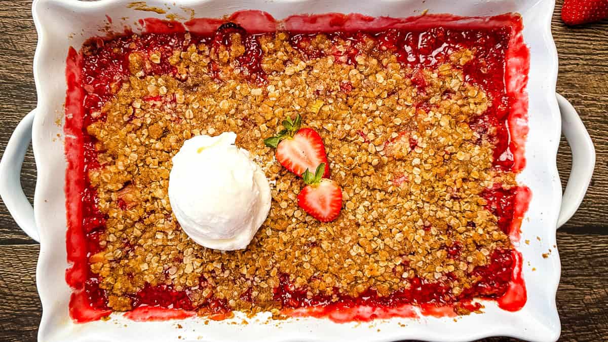A white baking dish of strawberry rhubarb crisp topped with a scoop of vanilla ice cream and fresh strawberries.