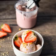 A bowl filled with two scoops of strawberry cheesecake ice cream garnished with fresh strawberries and crispy graham cracker crumbs.