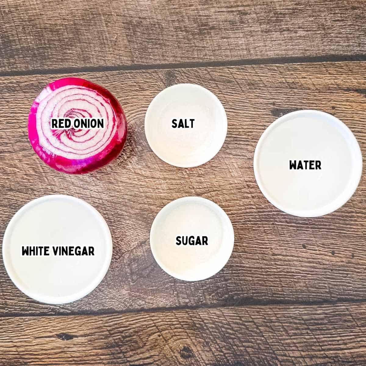Labeled ingredients for quick pickled red onions arranged on a wooden background, including a red onion, white vinegar, water, granulated sugar, and salt.