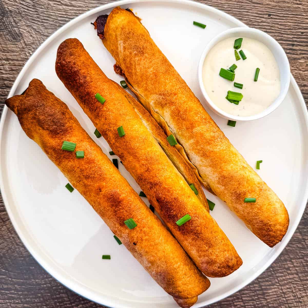 Three golden-brown baked pulled pork taquitos on a white plate, garnished with chopped green onions and served with ranch dipping sauce.