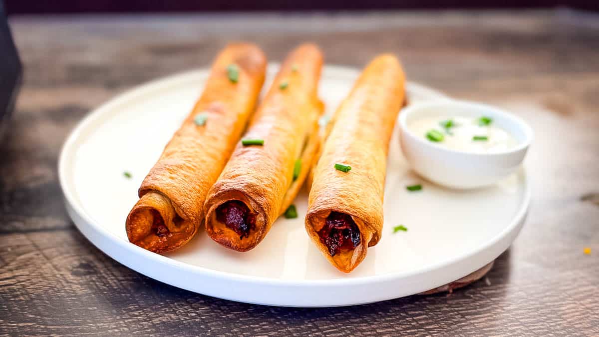 Three golden pulled pork taquitos on a white plate, with crispy edges.