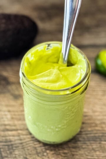 Fresh avocado lime crema in a glass jar with spoon, with avocado and lime in background on rustic wood surface.