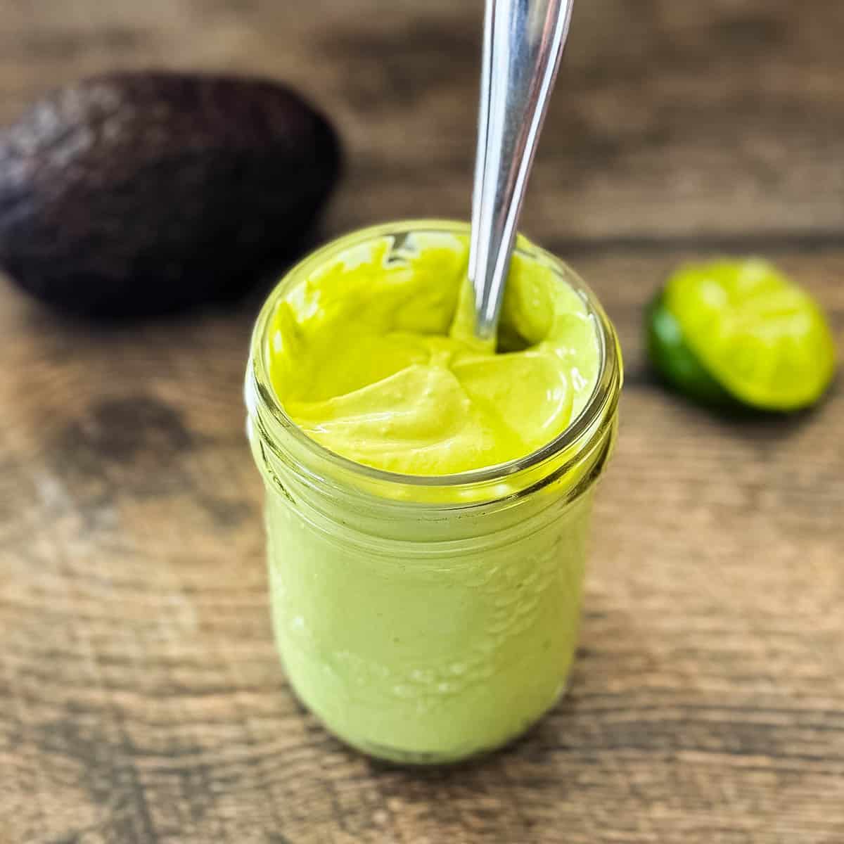 Fresh avocado lime crema in a glass jar with spoon, with avocado and lime in background on rustic wood surface.