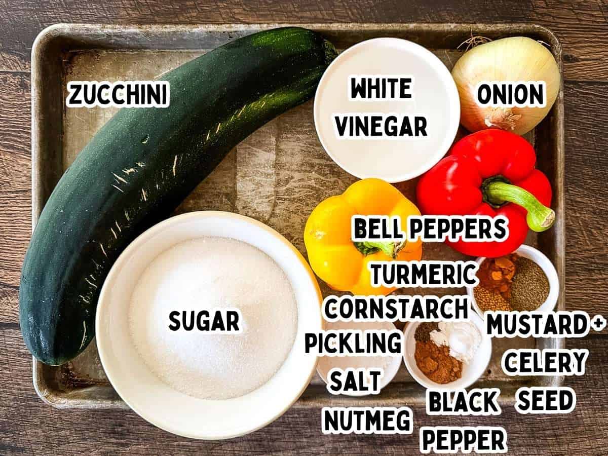 Labeled ingredients for zucchini relish arranged on a wooden table, including zucchini, onion, bell peppers, vinegar, sugar, and spices.