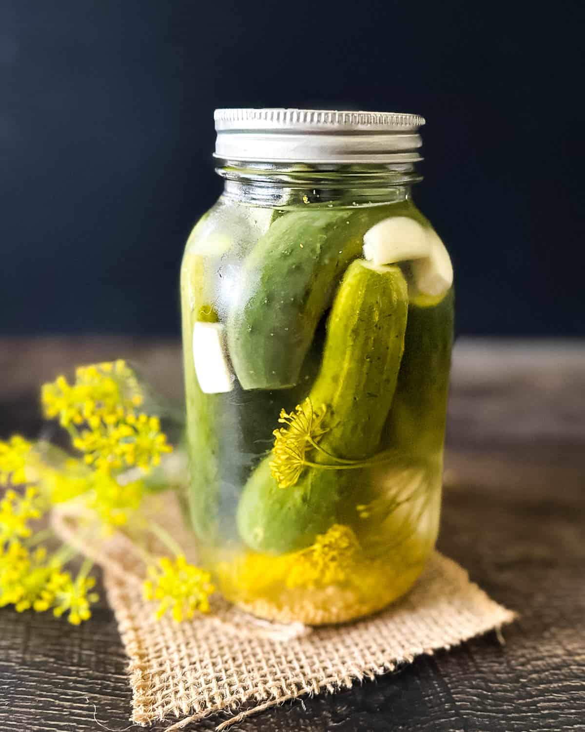 Fresh refrigerator dill pickles in a mason jar with garlic cloves and dill flowers, sitting on a rustic wooden table with burlap.