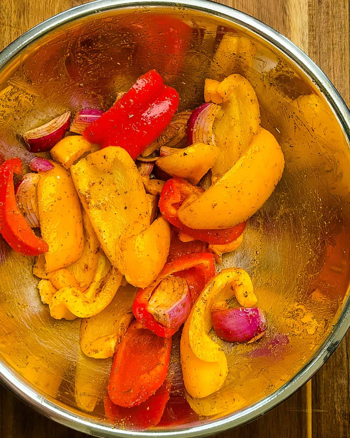 Sliced bell peppers and red onions tossed with olive oil and seasoning in a stainless steel mixing bowl.