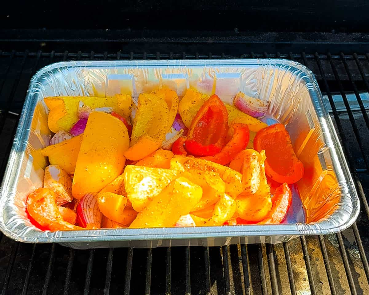 Foil pan filled with seasoned red onions and bell peppers placed on a pellet grill inside the smoker.