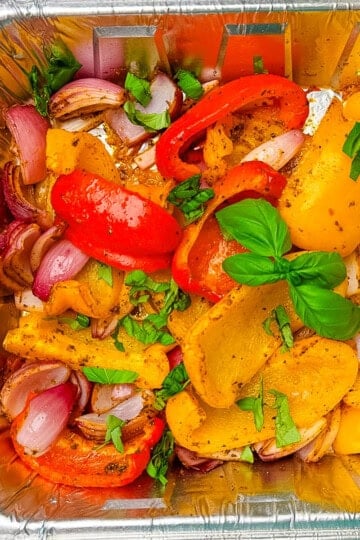 Smoked bell peppers and red onions in a disposable foil pan, seasoned and garnished with fresh chopped basil and whole basil leaves, ready to serve.