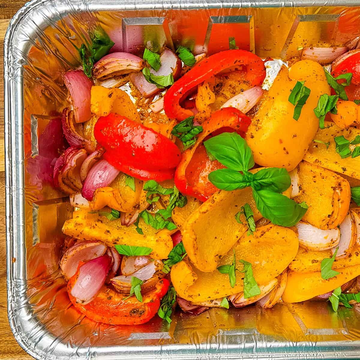 Smoked bell peppers and red onions in a disposable foil pan, seasoned and garnished with fresh chopped basil and whole basil leaves, ready to serve.
