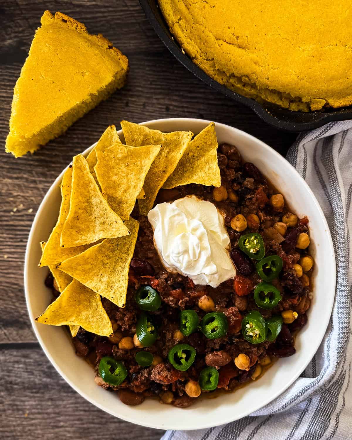 Cornbread slice served with a bowl of smoked chili topped with tortilla chips, sour cream, and jalapeno peppers.