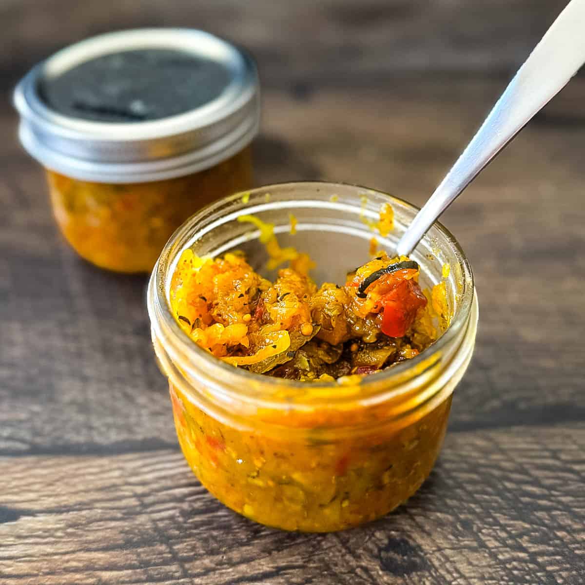 Glass jar filled with homemade zucchini relish on a wooden surface, with a spoon dipped inside.