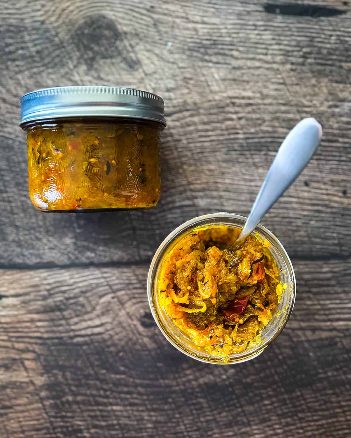 Open jar of homemade zucchini relish with a spoon next to a sealed jar, both on a rustic wood surface.