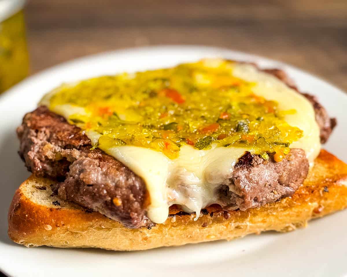 Open-faced cheeseburger on toasted bun topped with zucchini relish and melted cheese, served on a white plate.