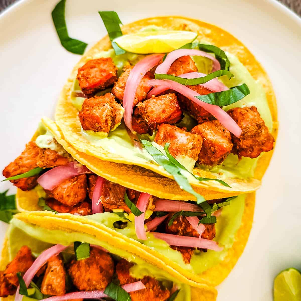 Three air fryer salmon tacos on corn tortillas topped with avocado lime crema, pickled red onions, and fresh cilantro, served with lime wedges.