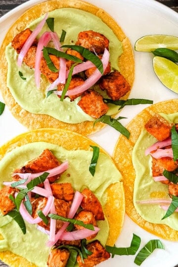 Air fryer salmon tacos on corn tortillas topped with avocado lime crema, pickled red onions, and fresh cilantro, served with lime wedges on a white plate.