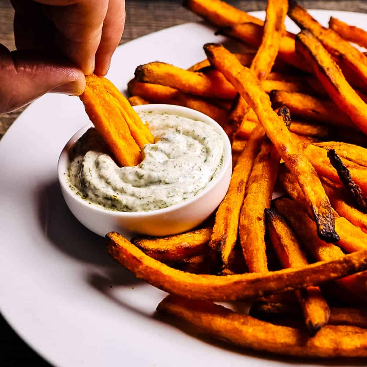 Hand dipping a sweet potato fry into a creamy homemade dill sauce on a white plate.
