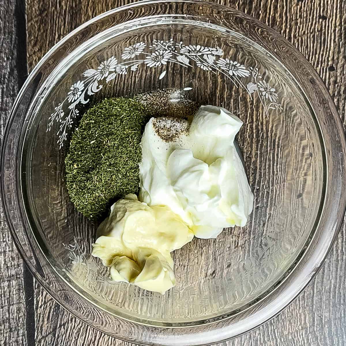 Glass mixing bowl with sour cream, mayonnaise, dried dill, salt, and pepper ready to make homemade dill mayo sauce.