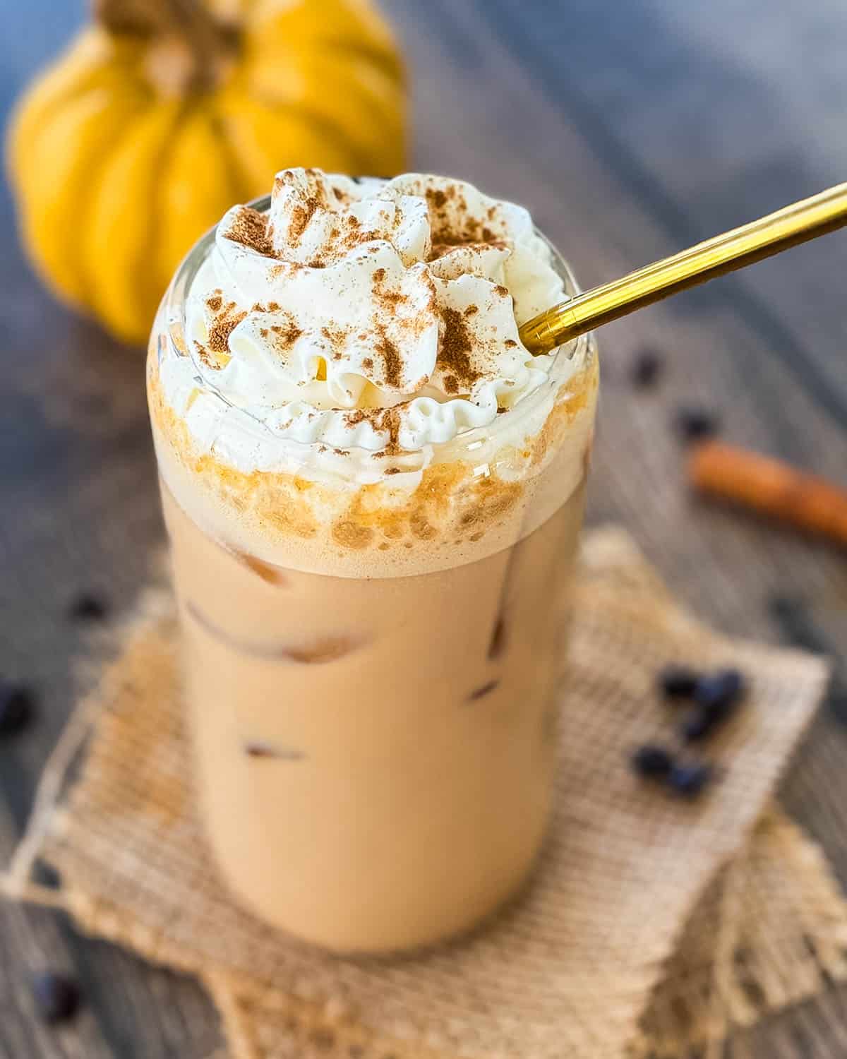 Finished iced pumpkin spice latte topped with whipped cream and a sprinkle of cinnamon, served with a gold straw in a tall glass.